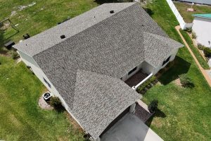 Aerial view of a single-story house with a gray shingle roof, driveway, and green lawn surrounding the home.