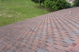 Angle view of a sloped roof covered in brown and gray asphalt shingles, with a grassy yard and trees in the background.