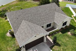 Aerial view of a beige, single-family home with a brown multi-gable shingle roof, attached two-car garage, and manicured lawn nearby water edge.