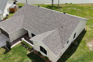 Aerial view of a beige single-story house with a brown shingle roof, U-shaped layout, front entry with small porch, and a green lawn nearby a body of water in the background.
