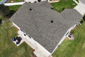 Aerial view of a single-story house with a multi-hipped gray shingle roof, surrounding lawn, and a small patio with a grill.