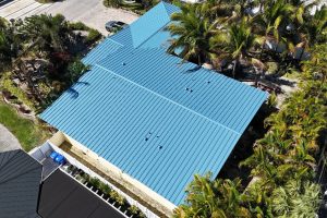Aerial view of a house with a bright blue metal roof surrounded by palm trees and greenery.
