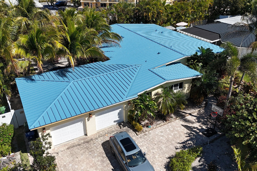 Aerial view of a house with a bright blue metal roof, surrounded by palm trees, and a driveway with a silver SUV and a basketball hoop nearby.