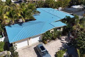Aerial view of a single-story house with a bright blue metal roof, palm trees surrounding the property, and a silver SUV parked in the driveway.