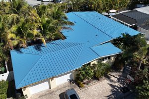 Aerial view of a house with a bright blue metal roof, three white garage doors, and a stone driveway surrounded by palm trees.