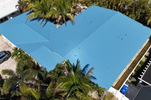Aerial view of a house with a bright blue metal roof, surrounded by palm trees; a car is parked in the driveway.