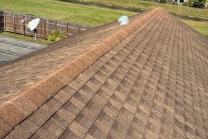 Brown asphalt shingle roof viewed from the ridge, with a fenced backyard and two satellite dishes in the background.