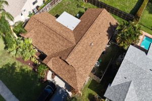 Aerial view of a brown shingle-roofed house with palm trees, a driveway, and a fenced backyard.