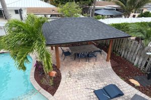 Backyard with a dark-shingled gazebo over a brick patio, a dining table and chairs, next to a swimming pool and palm tree.