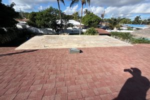 Overhead view of a red shingle roof with a light concrete flat section; a vent protrudes and a photographer's shadow appears in the foreground.