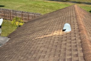 Brown shingle roof seen from above with a damaged curved vent and a lighter repair patch; fence and grass in the background.
