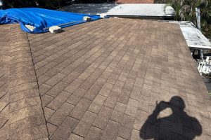 Brown shingle roof under repair with a blue tarp secured by concrete blocks; photographer's shadow is visible in the foreground.