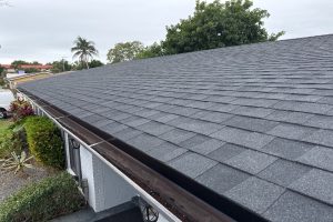 Side view of a gray shingle roof with a brown gutter along the edge of a white brick house, greenery in the foreground and a cloudy sky above.