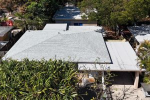 Aerial view of a single-story house with a light gray shingled roof, a carport on the right, and dense green shrubs in front.