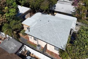 Aerial view of a single-story house with a light gray shingled hip roof, surrounded by trees and neighboring buildings.