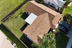 Aerial view of a suburban house with a brown shingle roof, green yard, and a driveway with a covered car beside palm trees.