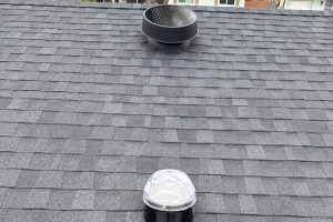 Round roof vent with rain cap on a gray shingled roof, viewed from above, with a translucent dome light on a black base in the foreground