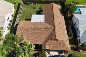 Aerial view of a brown shingle-roof house with a complex roofline, surrounded by a fenced yard and palm trees; neighboring homes and a pool are visible nearby.