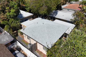 Aerial view of a beige single-story house with a light gray metal roof, surrounded by trees and nearby homes.