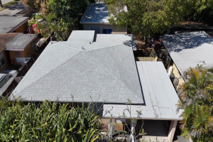 Aerial view of gray shingled roof with triangular ridge lines, surrounded by trees and neighboring houses, seen from above.