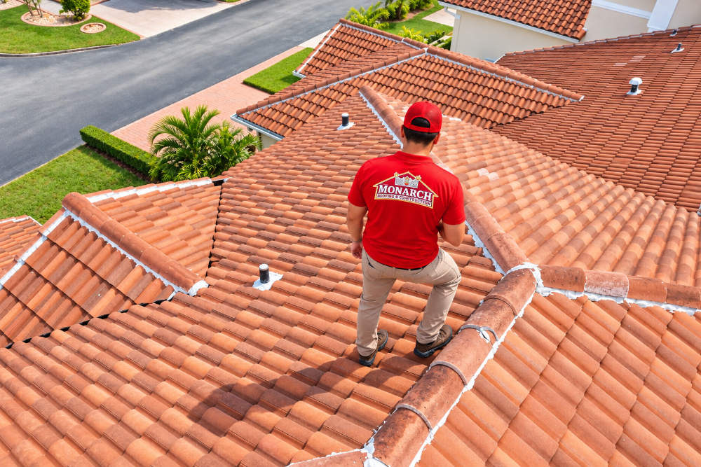 Roofer in a red Monarch Roofing shirt stands on a terracotta-tiled roof in a suburban neighborhood.